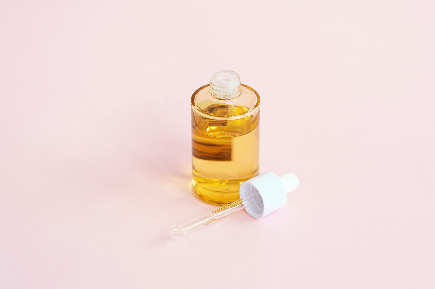Glass dropper bottle of amber serum with its dropper lying beside it on a pale pink surface